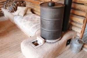 A rustic indoor scene featuring a large black metal barrel stove on a clay bench, with a patterned blanket and pillows on the bench. Stacked firewood and a metal bucket are nearby.