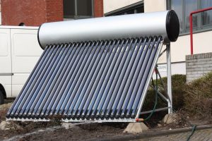 A solar water heater with multiple vacuum tubes arranged in a tilted frame, positioned outdoors near a building. The system has a cylindrical storage tank at the top and hoses connected at the base.