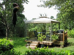 A gazebo in a lush garden with a wooden deck, green chairs, and a barbecue grill under a canopy.