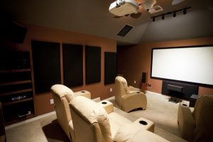 A home theatre with beige recliner seats, a large projection screen, wall-mounted acoustic panels, and a ceiling projector.