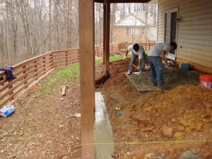 Two people are working on a concrete foundation next to a house, with a wooden fence and trees in the background.