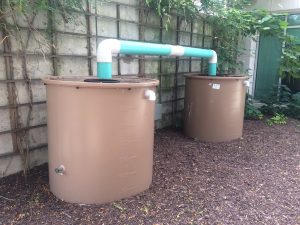 Two large brown cylindrical tanks connected by a white and blue pipe, set outdoors against a wall covered with plants. The tanks have measurement markings on them and are placed on mulch-covered ground.